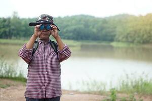 ásia Senior mulher vai viajando sozinho, usar binocular para pesquisa natureza às a lago. conceito, lazer atividade. Senior estilo de vida. gastar Tempo para viagem depois de aposentado. foto