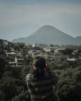 Individual dentro traseiro visualizar, cercado de panorama e montanhas adulto admirando montanha alcance com edifícios, multidão, e azul céu. foto