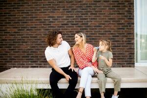 família com uma mãe, pai e filha sentado lado de fora em a passos do uma frente varanda do uma tijolo casa foto