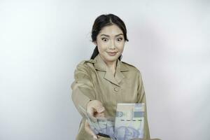 uma sorridente jovem governo trabalhador mulher é vestindo cáqui uniforme segurando dinheiro dinheiro dentro indonésio rupia isolado de branco fundo foto