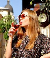 jovem de óculos escuros bebendo um copo de vinho tinto rosa ao ar livre em um café, cabelo loiro comprido vestido escuro foto