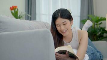 lindo ásia jovem mulher lendo livro em sofá às lar. foto