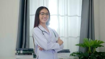 ásia mulher médico em pé com braços cruzado é vestindo uniforme branco roupão e estetoscópio dentro exame quarto às a clínica. foto