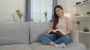 lindo ásia jovem mulher lendo livro em sofá às lar. foto