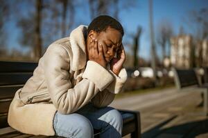 ao ar livre retrato do depressivo afro-americano mulher em ensolarado dia. ela é sentado em Banco dentro a rua. foto