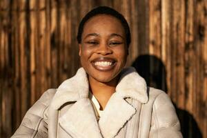 ao ar livre retrato do feliz afro-americano mulher. ela é em pé dentro frente do de madeira parede e sorridente. foto