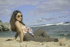 jovem ásia mulher sentar em a de praia areia. retrato sexy ásia senhora viajando e relaxante dentro a verão com tropical natureza. foto