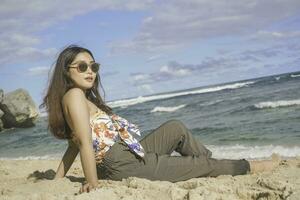 jovem ásia mulher sentar em a de praia areia. retrato sexy ásia senhora viajando e relaxante dentro a verão com tropical natureza. foto