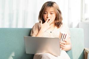 jovem asiática sentada no sofá, comendo e assistindo filmes no laptop