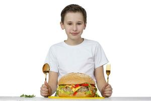 jovem Garoto com grande Hamburger isolado dentro branco. uma adolescente detém uma dourado garfo e uma colher. foto