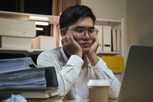 ásia jovem cansado funcionários homem de negocios usando Área de Trabalho computador tendo excesso de trabalho projeto durante a noite dentro escritório. foto