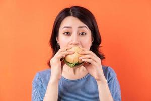 jovem mulher asiática comendo hambúrguer em fundo laranja foto