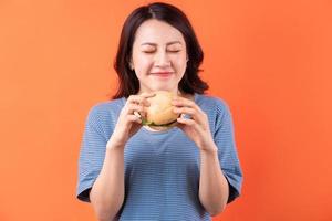 jovem mulher asiática comendo hambúrguer em fundo laranja foto