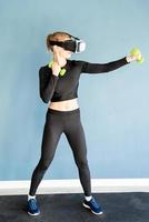 jovem loira com roupas esportivas e óculos de realidade virtual em pé na esteira de ginástica, malhando com dubbells