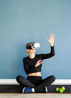 jovem loira com roupas esportivas e óculos de realidade virtual, sentada no tapete de fitness usando o menu interativo de VR foto