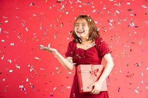 jovem asiática em um vestido segurando uma caixa de presente vermelha com uma expressão alegre no fundo foto