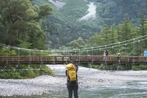caminhante com mochilas com kappa ponte Kamikochi com rio e okuhotaka dia montanha fundo. Kamikochi famoso Lugar, colocar para caminhada e caminhada dentro matsumoto, Nagano, Japão foto