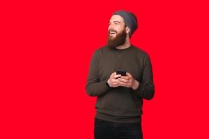 Homem barbudo hippie sorridente usando smartphone e olhando para longe sobre fundo vermelho