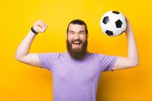homem espantado com barba gritando e comemorando a vitória, torcedor apoiando o time favorito e segurando uma bola de futebol foto