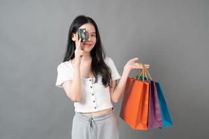 jovem mulher asiática segurando uma sacola de compras e um cartão de crédito em fundo cinza