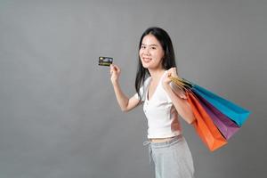 jovem mulher asiática segurando uma sacola de compras e um cartão de crédito em fundo cinza foto