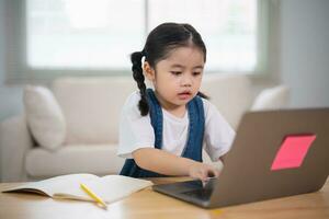 ásia bebê menina sorridente usar computador portátil ou escrevendo desenhando cor em Nota livro estude conectados em madeira mesa escrivaninha dentro vivo quarto às lar. Educação Aprendendo conectados a partir de casa conceito. foto