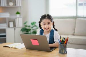 ásia bebê menina sorridente usar computador portátil ou escrevendo desenhando cor em Nota livro estude conectados em madeira mesa escrivaninha dentro vivo quarto às lar. Educação Aprendendo conectados a partir de casa conceito. foto