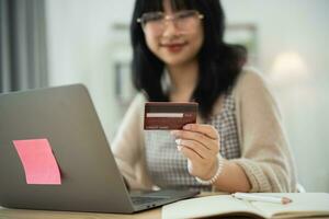 mulher segurando mostrando o cartão de crédito para fazer compras online. mulher asiática trabalhando em casa. compras on-line, e-commerce, internet banking, gastar dinheiro, trabalhar em casa. foto