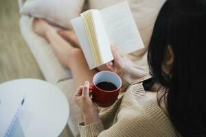 alegre ásia mulher lendo livro e beber quente café enquanto sentado em sofá sofá dentro vivo quarto às lar. ásia mulheres relex lendo livro sorrir às sofá dentro a casa. atividade passatempo às casa conceito. foto