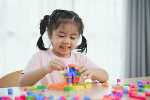alegre ásia menina feliz e sorridente jogando colorida Lego brinquedos, sentado em a mesa dentro a vivo sala, criativamente jogando com Lego, construção colorida estruturas criatividade Imagine. foto