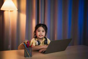 ásia menina sorridente, rindo, excitado, olhando, vestindo branco fone de ouvido e usando computador portátil transmissão noite luz dentro vivo quarto às lar. fofa menina assistindo videos enquanto televisão Internet vício conceito. foto