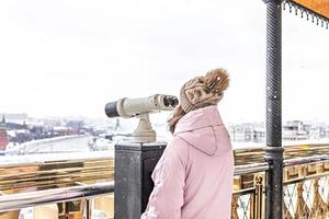uma jovem olha através de binóculos operados por moedas no deck de observação com vista para a cidade de uma altura ao pôr do sol. inverno, queda de neve