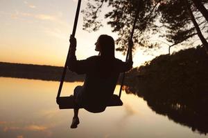 jovem romântica em um balanço sobre o lago ao pôr do sol. jovem viajante sentada no balanço na bela natureza, vista para o lago foto