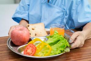 paciente asiático sênior ou idosa idosa comendo alimentos saudáveis de vegetais de café da manhã com esperança e feliz enquanto está sentado e com fome na cama no hospital. foto