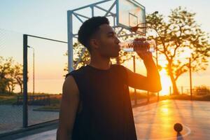 Preto homem fazendo Esportes dentro manhã, bebendo água em basquetebol quadra em nascer do sol foto