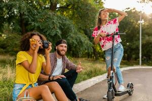 feliz jovem companhia do sorridente amigos sentado dentro parque em Relva com elétrico pontapé lambreta foto