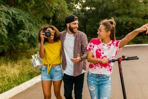 feliz jovem companhia do sorridente amigos caminhando dentro parque com elétrico pontapé lambreta foto