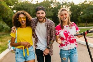 feliz jovem companhia do sorridente amigos caminhando dentro parque com elétrico pontapé lambreta foto