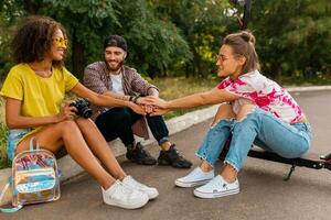 feliz jovem companhia do sorridente amigos sentado dentro parque em Relva com elétrico pontapé lambreta foto