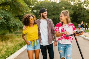 feliz jovem companhia do sorridente amigos caminhando dentro parque com elétrico pontapé lambreta foto