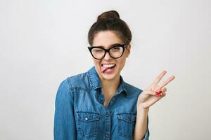 hipster jovem à moda mulher dentro na moda óculos e jeans camisa fazer engraçado face expressão, mostrando língua, piscando, Paz placa gesto, isolado em branco estúdio fundo, positivo foto