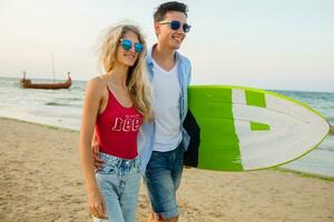 jovem sorridente casal tendo Diversão em de praia caminhando com surfar borda foto