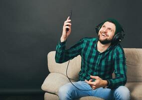 jovem hipster bonito barbudo homem sentado em uma sofá às lar, ouvindo para música foto