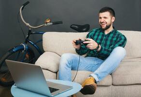 jovem hipster bonito barbudo homem sentado em sofá às lar, jogando vídeo jogos foto