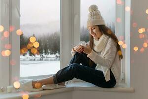 jovem atraente mulher dentro à moda branco tricotado suéter, cachecol e chapéu sentado às casa em peitoril da janela às Natal segurando vidro neve bola presente decoração, inverno floresta visualizar, luzes bokeh foto