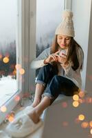 jovem atraente mulher dentro à moda branco tricotado suéter, cachecol e chapéu sentado às casa em peitoril da janela às Natal segurando vidro neve bola presente decoração, inverno floresta visualizar, luzes bokeh foto