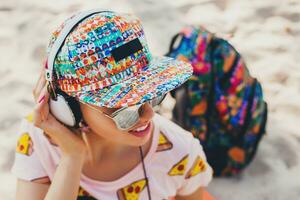 atraente hipster mulher sentado em de praia ouvindo para música em fones de ouvido foto