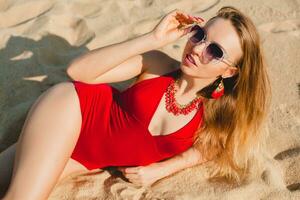 jovem lindo loiro mulher banhos de sol em areia de praia dentro vermelho natação terno, oculos de sol foto