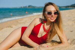 jovem lindo loiro mulher banhos de sol em areia de praia dentro vermelho natação terno, oculos de sol foto