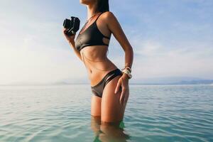 lindo mulher, bronzeada pele, Preto bikini roupa de banho, em pé dentro azul água foto